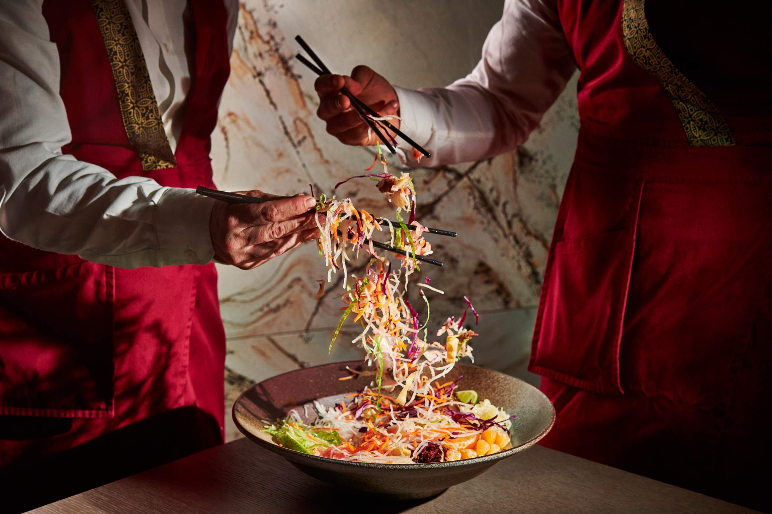 Two people use chopsticks to toss and mix a salad