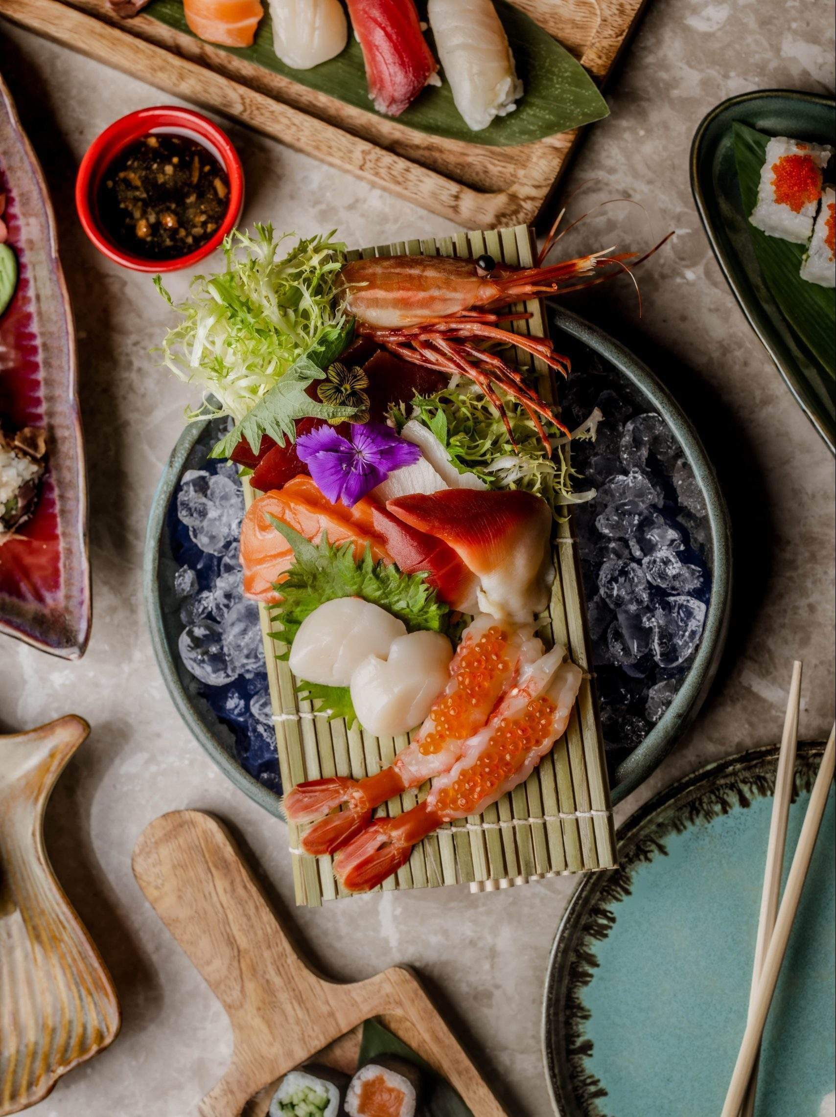 A dish with sashimi, a shrimp, salad leaves and flowers on a plate with ice on a table at a restaurant