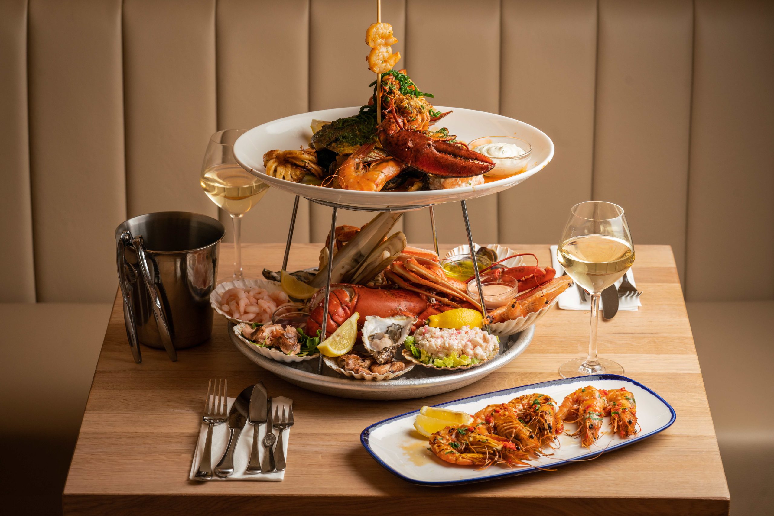 A two-tier seafood platter filled with shrimps, clams and oysters, with a small dish of shrimps, cutlery and a wine glass on a restaurant table