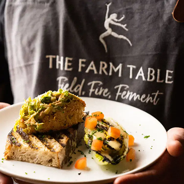 A waiter wearing an apron that says 'The Farm Table' holds a white plate with a toast topped with a scoop of terrine at The Farm Table, one of the most sustainable restaurants in the UK