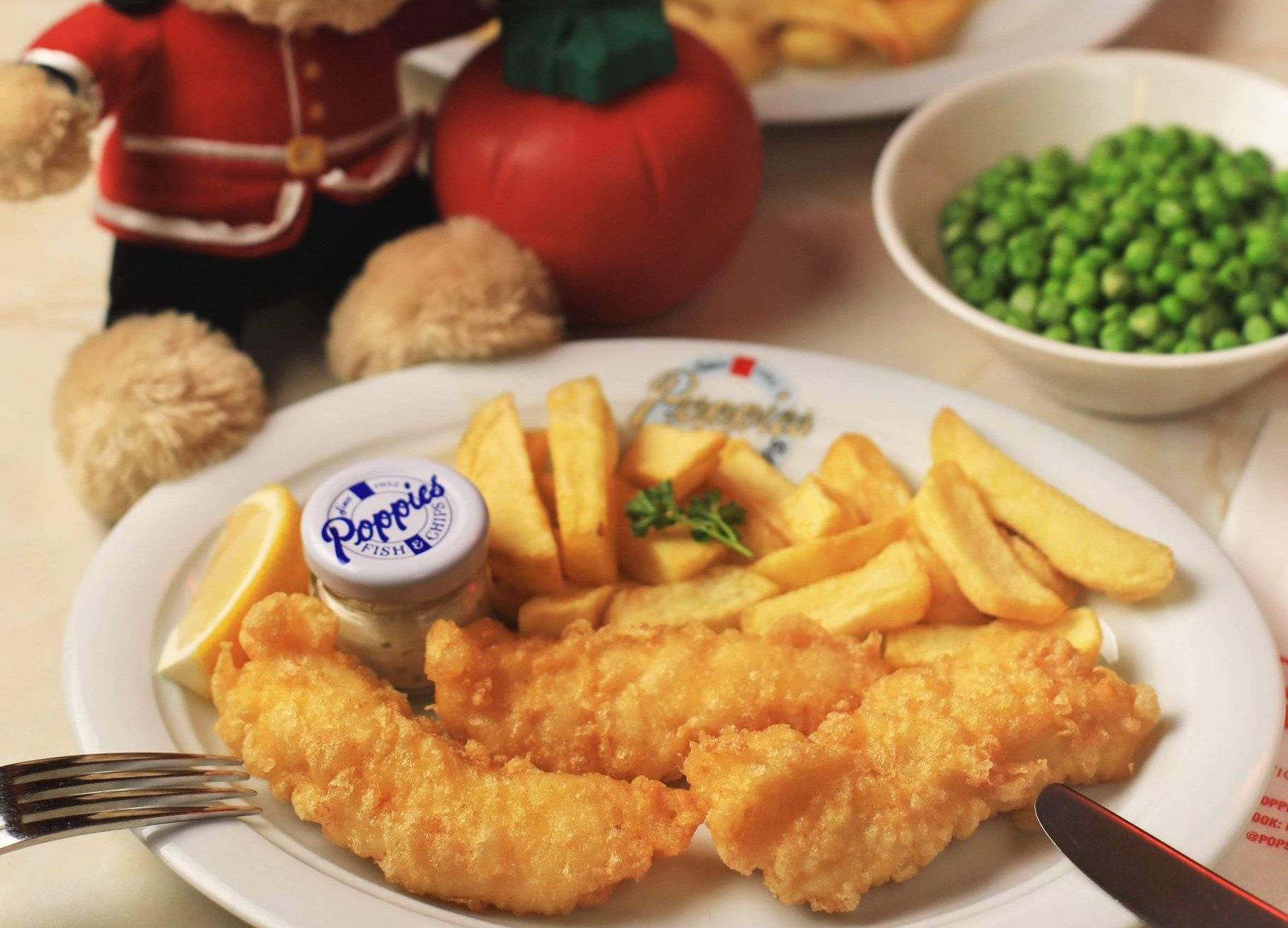 A plate of fish and chips with garden peas and a plush teddy bear wearing a guard costume at Poppies.