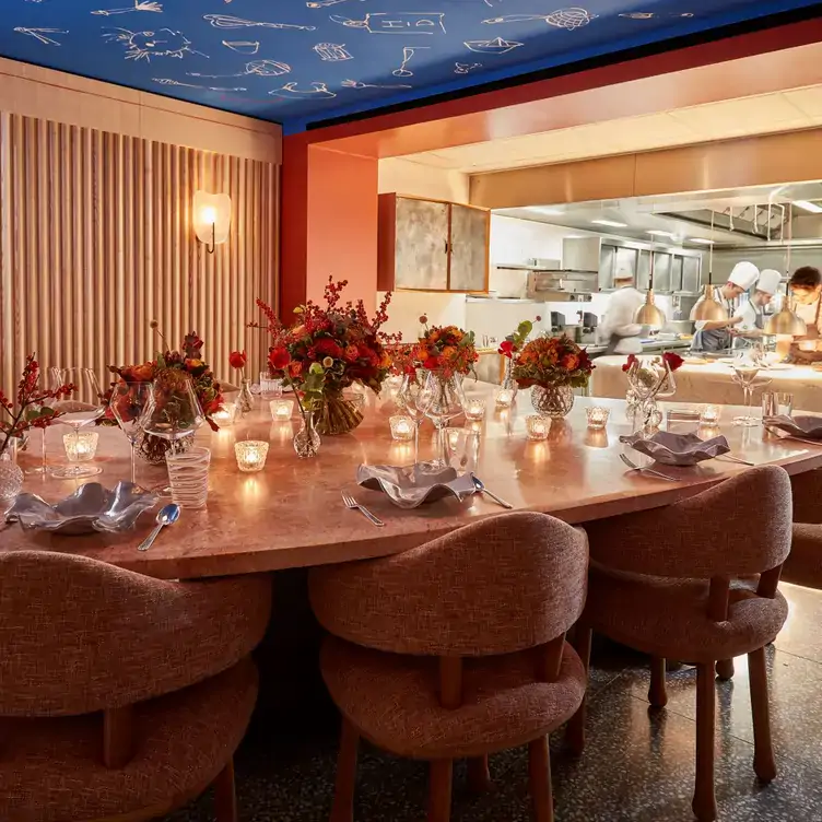 A round wooden table adorned with flowers and candles with an open-kitchen in the background at Hélène Darroze at The Connaught