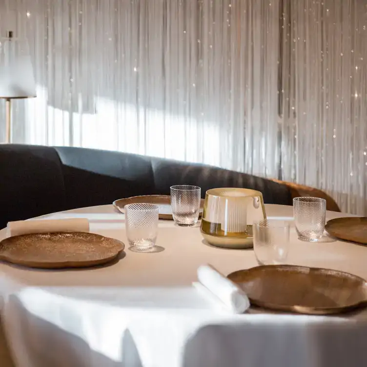 A table with white tablecloth and elegant plates, glasses and cutlery sits in front of a shimmering curtain at Alain Ducasse at The Dorchester, a restaurant in London