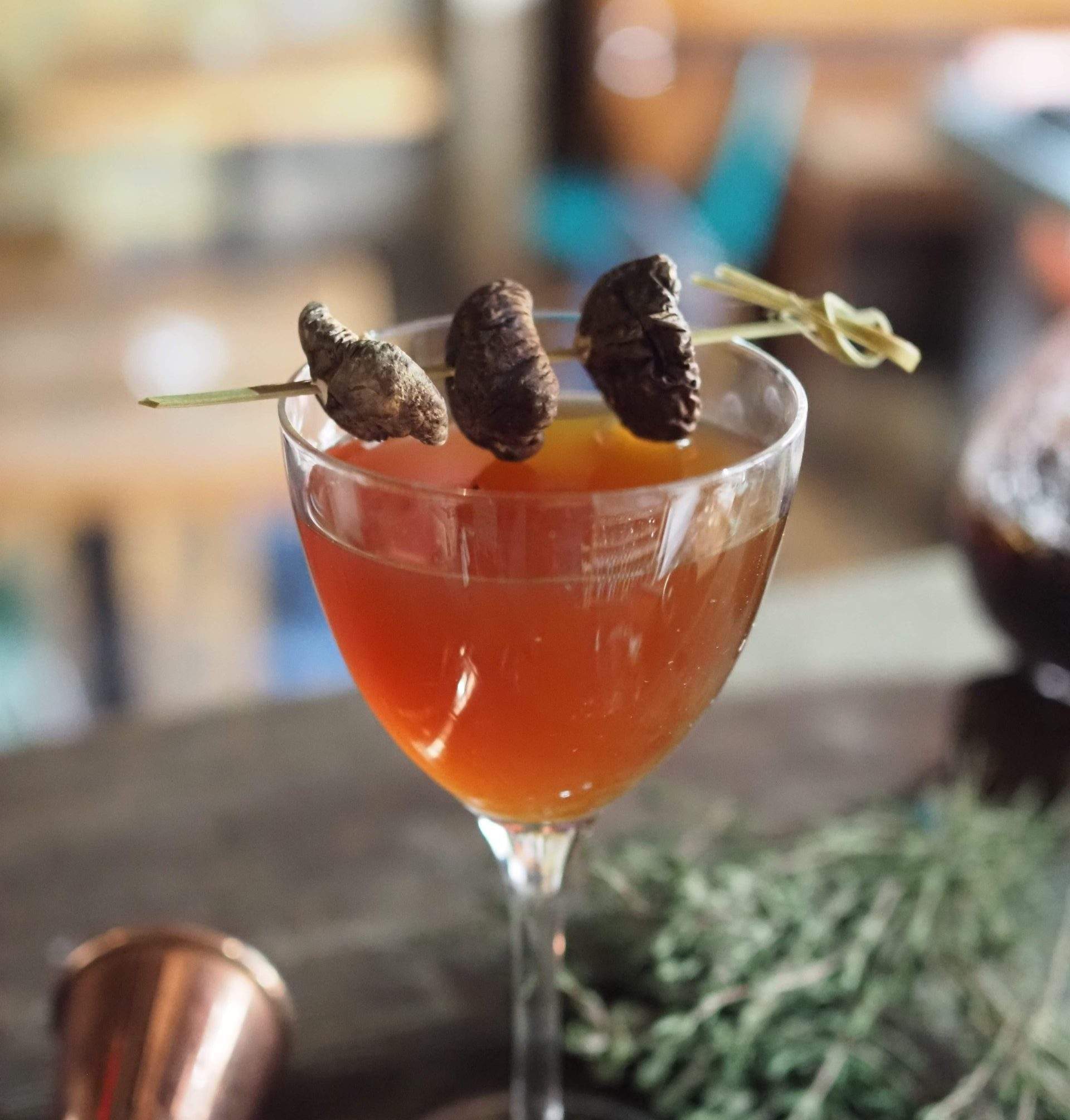 An orange cocktail topped with a skewer featuring three brown mushrooms on a dark wooden table from Love Shack in London.