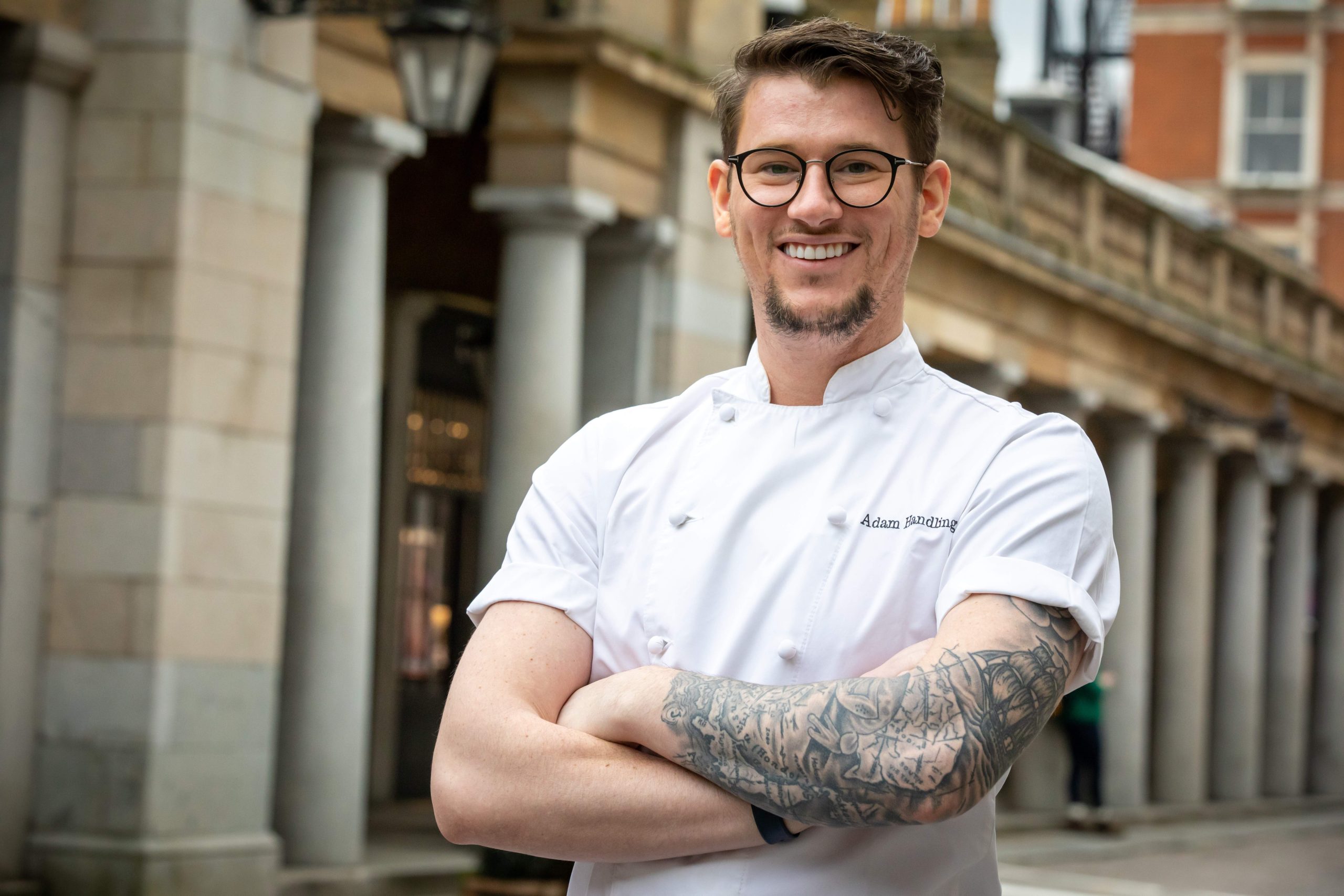 A headshot of chef Adam Handling wearing a white chef’s uniform and smiling in the streets of Covent Garden in London.