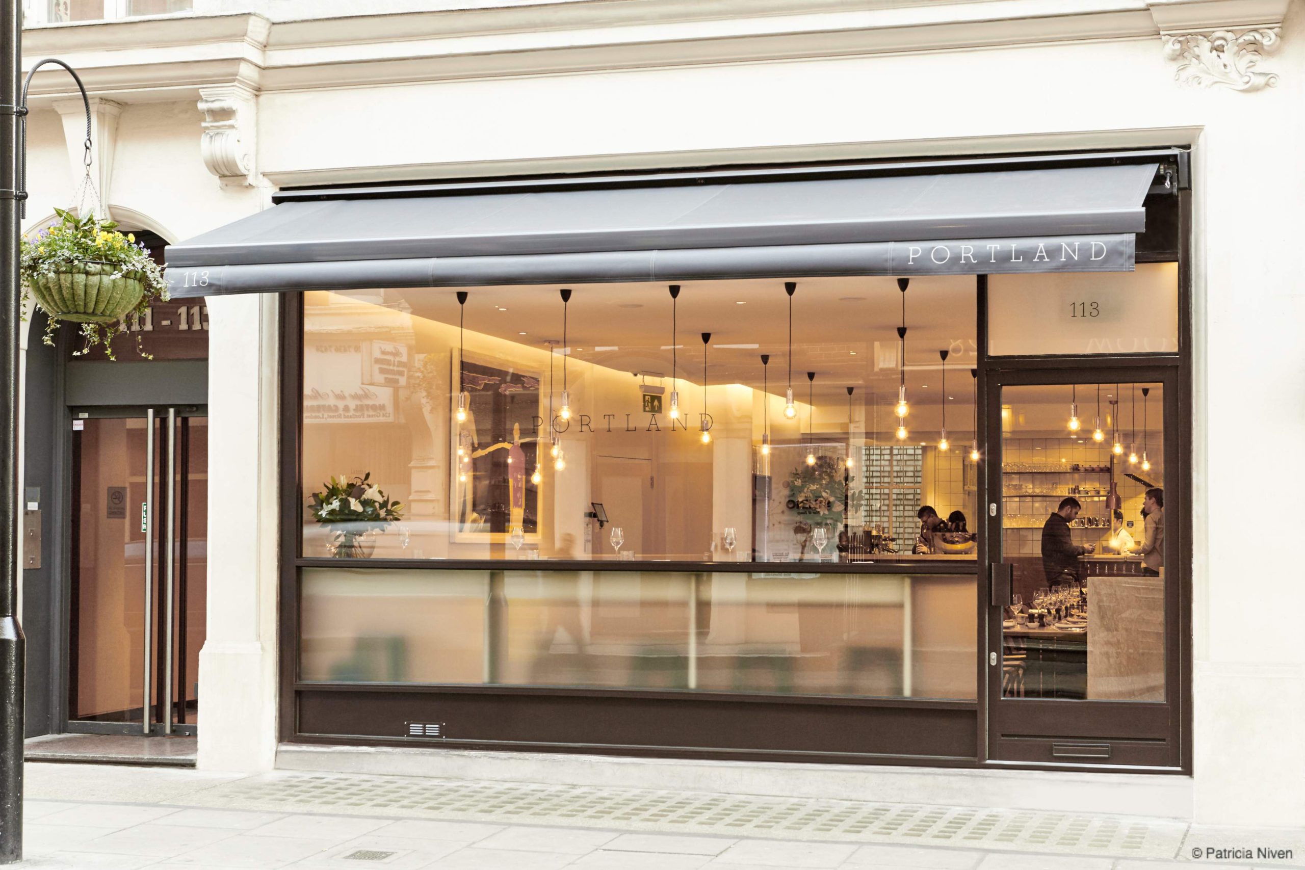 The white facade of Portland: a bright restaurant featuring a windowed wall revealing cosy interiors with hanging lights.