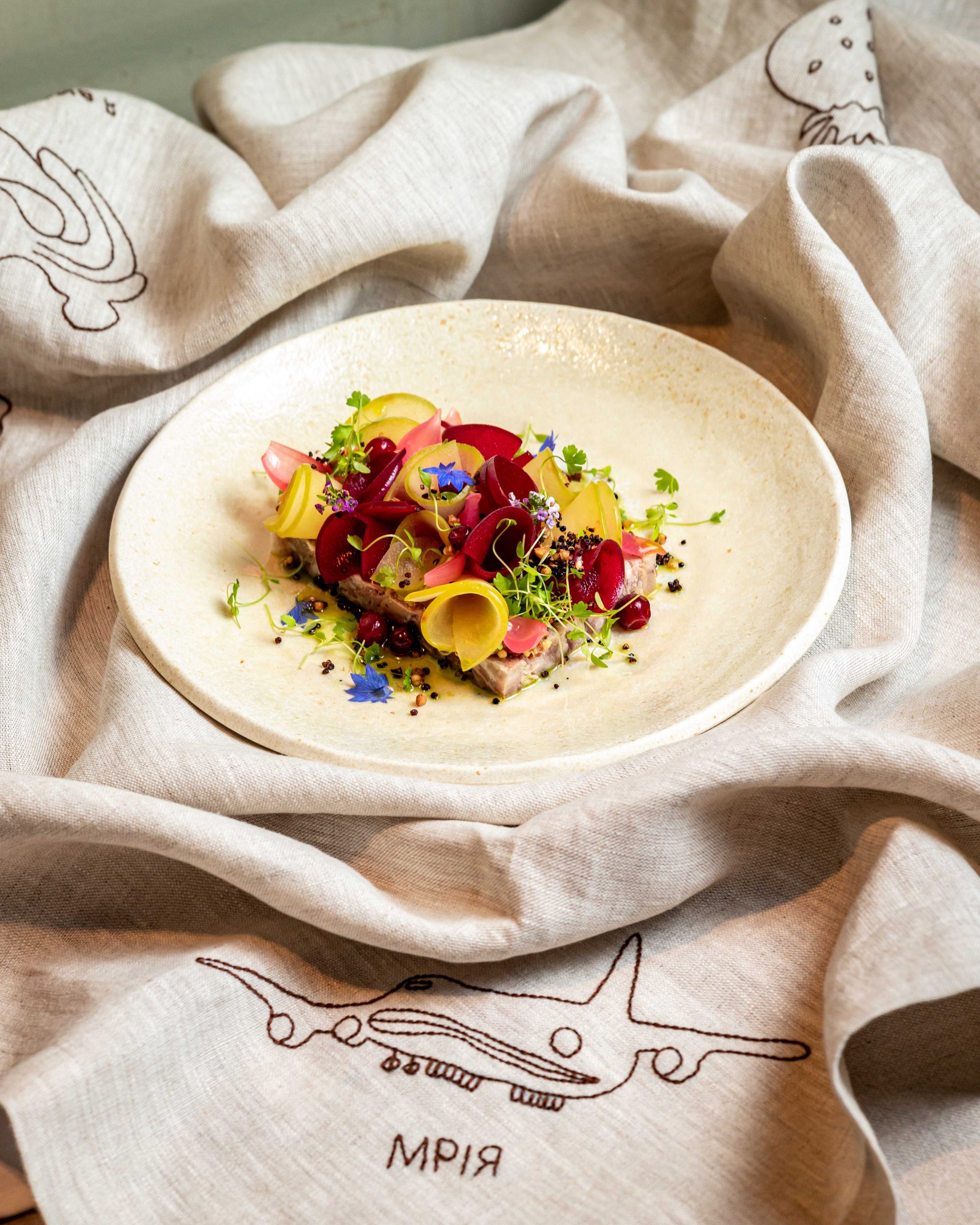 A white dish filled with greens, flowers, and pickles sits on top of a tablecloth embroidered with the aircraft Mriya at the restaurant of the same time in London