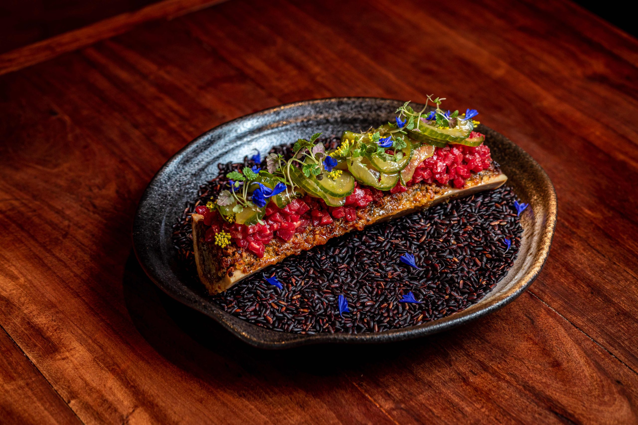 A roasted bone marrow sits on top of a plate filled with black rice, garnished with beef tartare and spicy pickled cucumbers at Mriya, a restaurant in London.