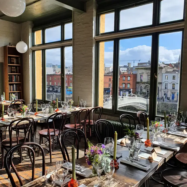 The interior of the Winding Stair, showing long tables decorated with flowers and tall windows looking out to the city with a view of the Ha’Penny Bridge.