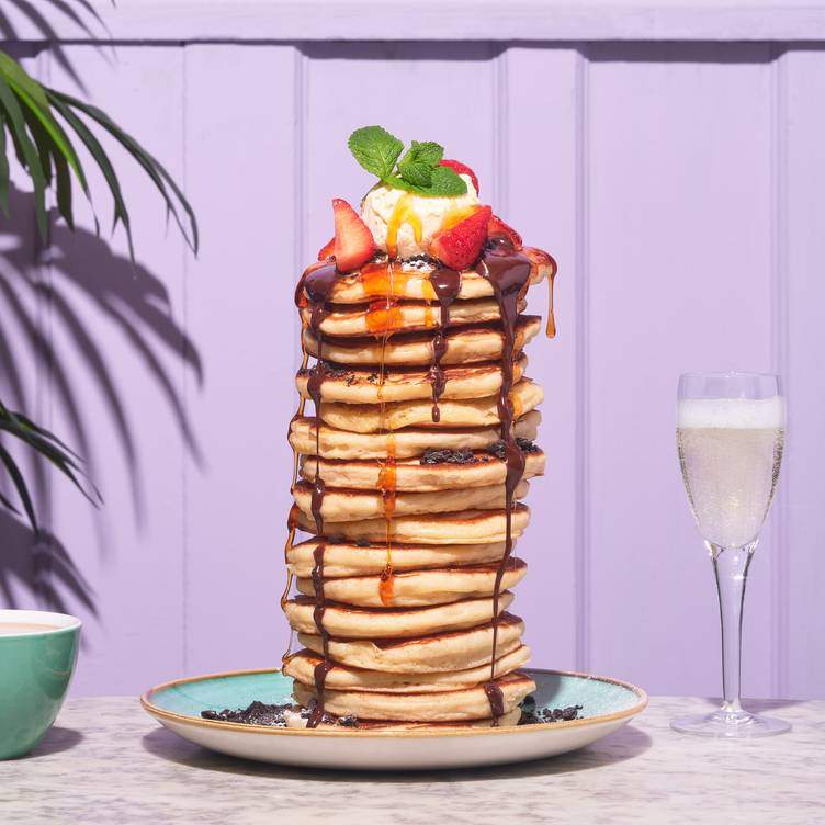 A stack of pancakes with chocolate sauce, ice cream and fruit with a glass of prosecco.