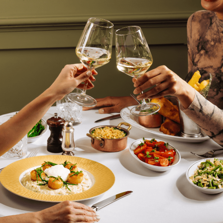 A couple of diners clink wine glasses in front of main dishes at some side dishes at the Ivy Buchanan Street Glasgow, one of the best brunch restaurants in Glasgow.