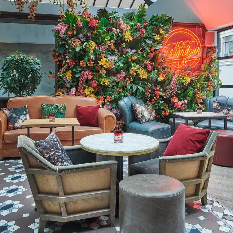 A colourful seating arrangement around tables in front of a floral display on the wall at 100 Wardour Street Drinks, one of the best live music restaurants in London.