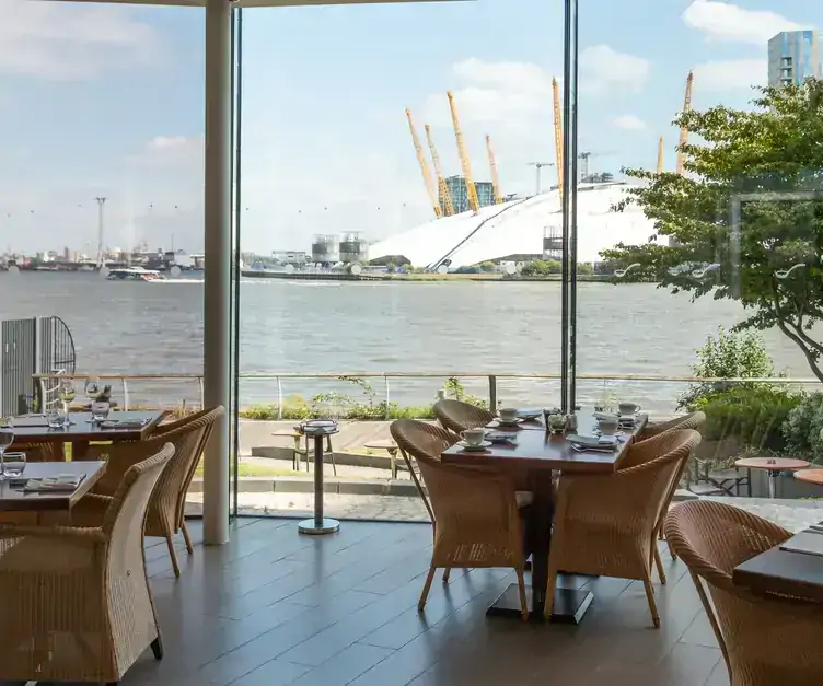 Tables and chairs near the tall windows at Scoff & Banter showing the view across the Thames and towards the O2.