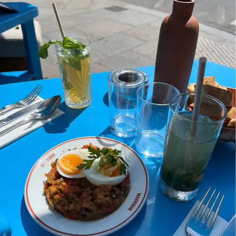 A bright blue outdoor table with two different cocktails and a dish topped with soft boiled eggs served at Mabrouk, one of Paris’ best restaurants.