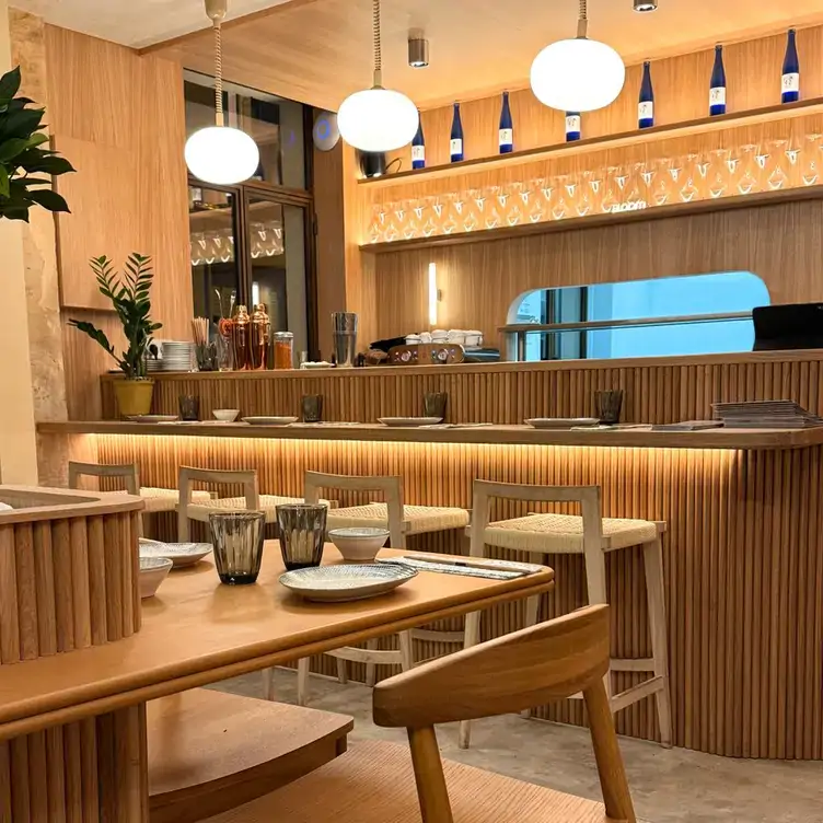 The pale brown interior of Bloom Sushi, showing the wooden counter and tables set with plates, chopsticks and glasses.