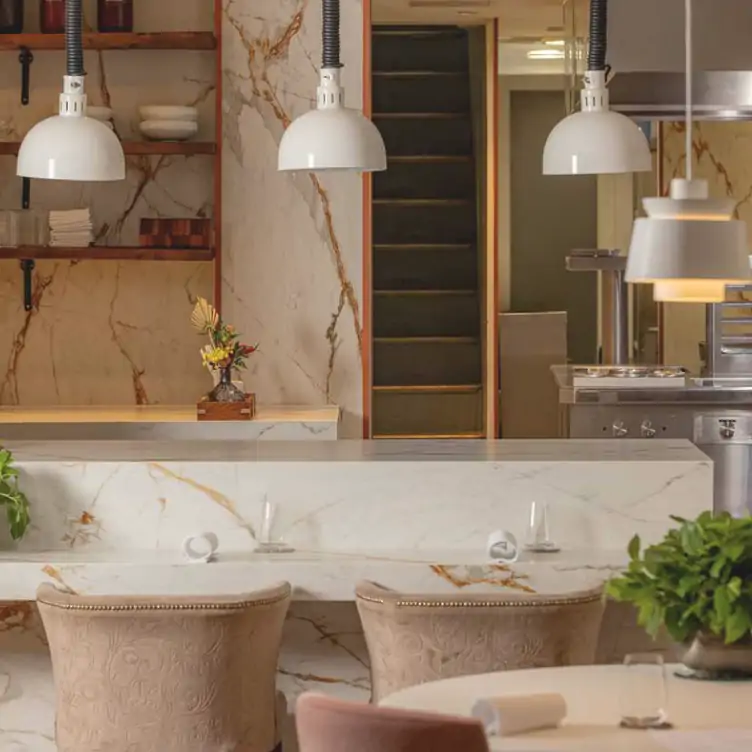 The interior of Frog by Adam Handling, showing the pale pink and beige chairs around tables and at the counter in front of the open kitchen.