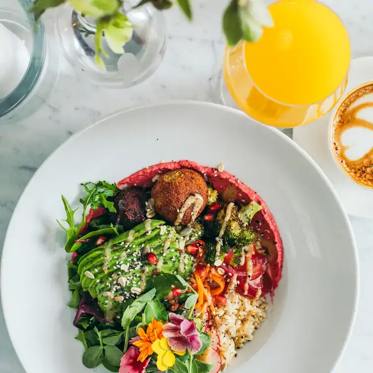 A dish of mixed vegan food including sliced avocado, with edible flower garnish and a glass of orange juice from Medicine Bakery.