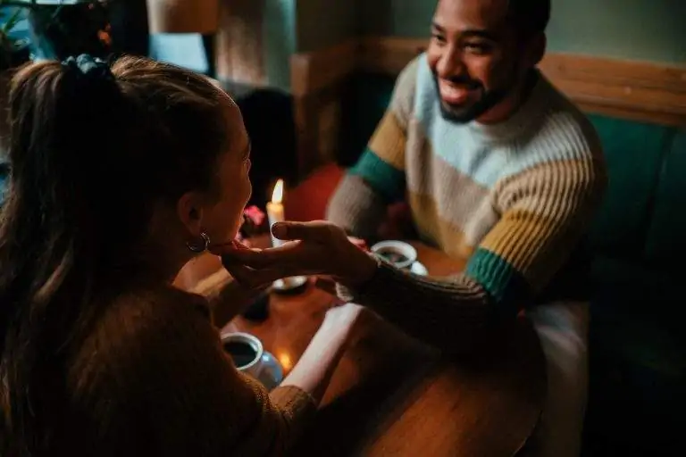 A couple sitting in front of each other at a restaurant table, the man touching the woman's face, with coffee and a candle