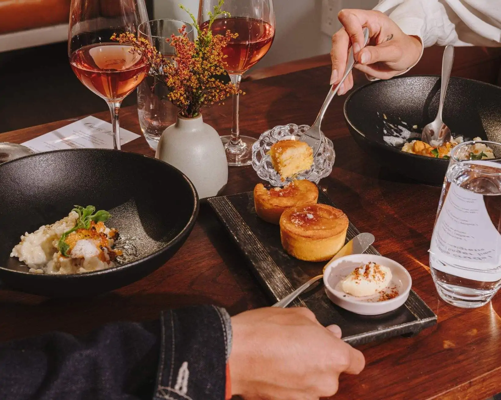 A dining table at a restaurant with a close up of a couple's hands, with dishes, dessert and two glasses of wine