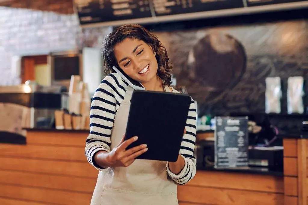 A waitress wearing a striped shirt and an apron speaks on the phone while looking at her tablet at a restaurant