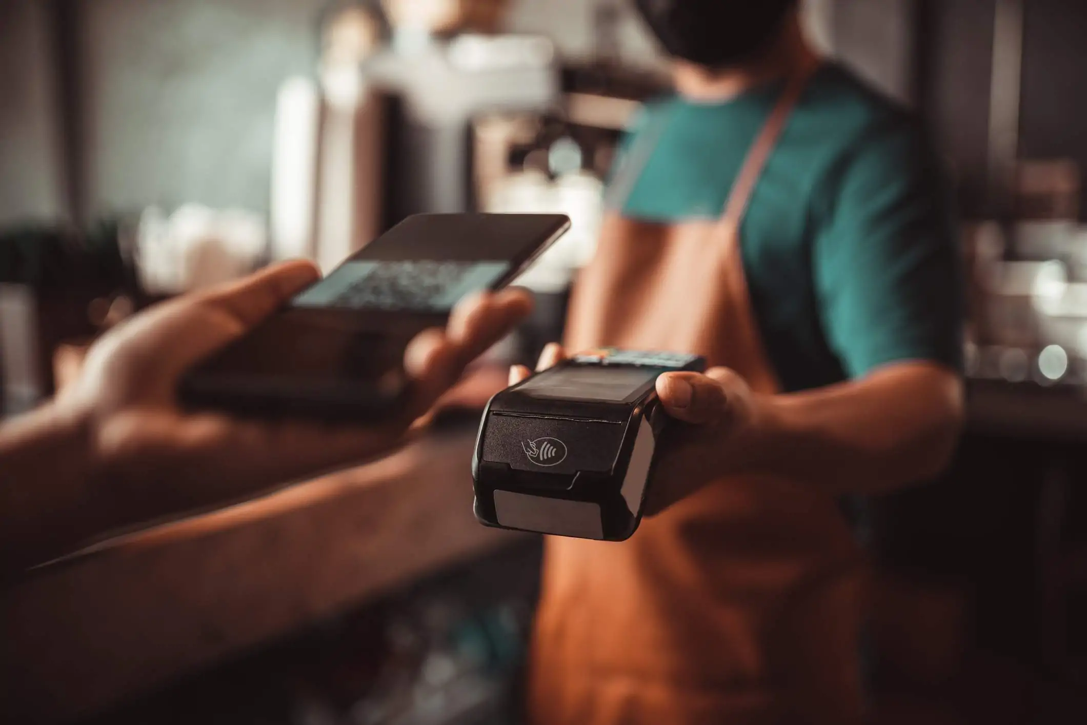 Image depicts a server wearing a green t-shirt and a brown apron holding a point-of-sales (POS) system out to a customer. The customer is holding their phone up to pay for their meal.